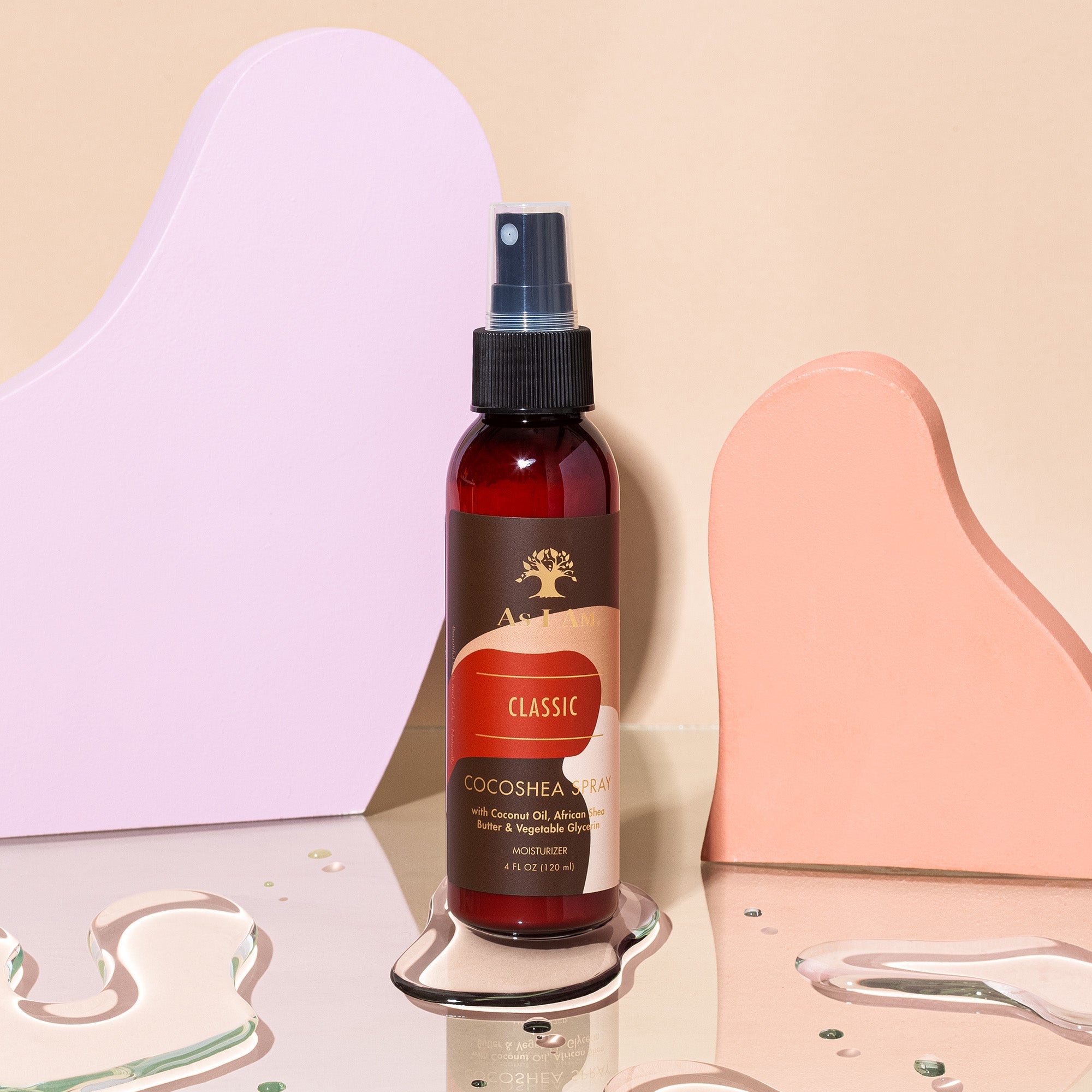 En brun sprayflaske med en hvid etiket står på en pastelfarvet baggrund. Flasken indeholder As I Am Classic CocoShea Spray og har et billede af et træ på etiketten. Produktet er designet til at give fugt og glans til håret.