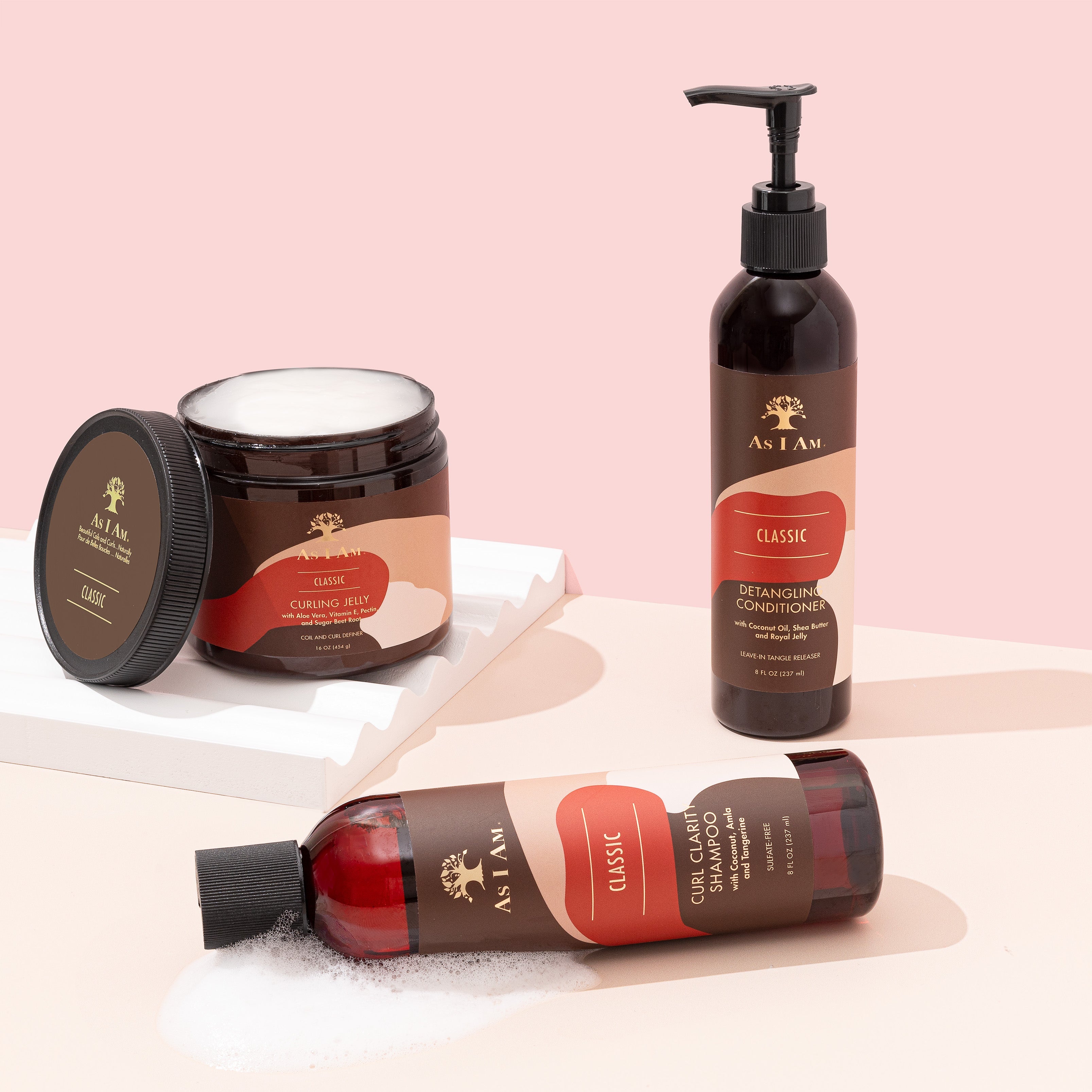 Tre hårplejeprodukter fra As I Am står på en lys bordplade. Fra venstre mod højre ses en mindre flaske shampoo, en stor krukke med hårcreme og en høj flaske med balsam. Produkterne har alle en brun farve og en hvid etiket med gulddetaljer.