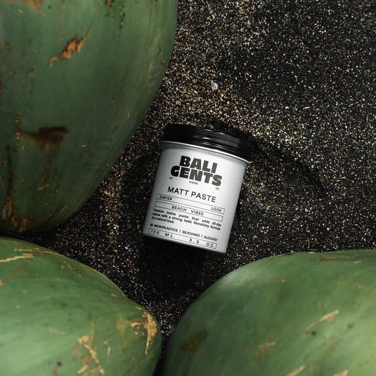 Bali Gents Matt Paste 100ml placed on a black sand beach surrounded by green coconuts gives a natural beach vibe.