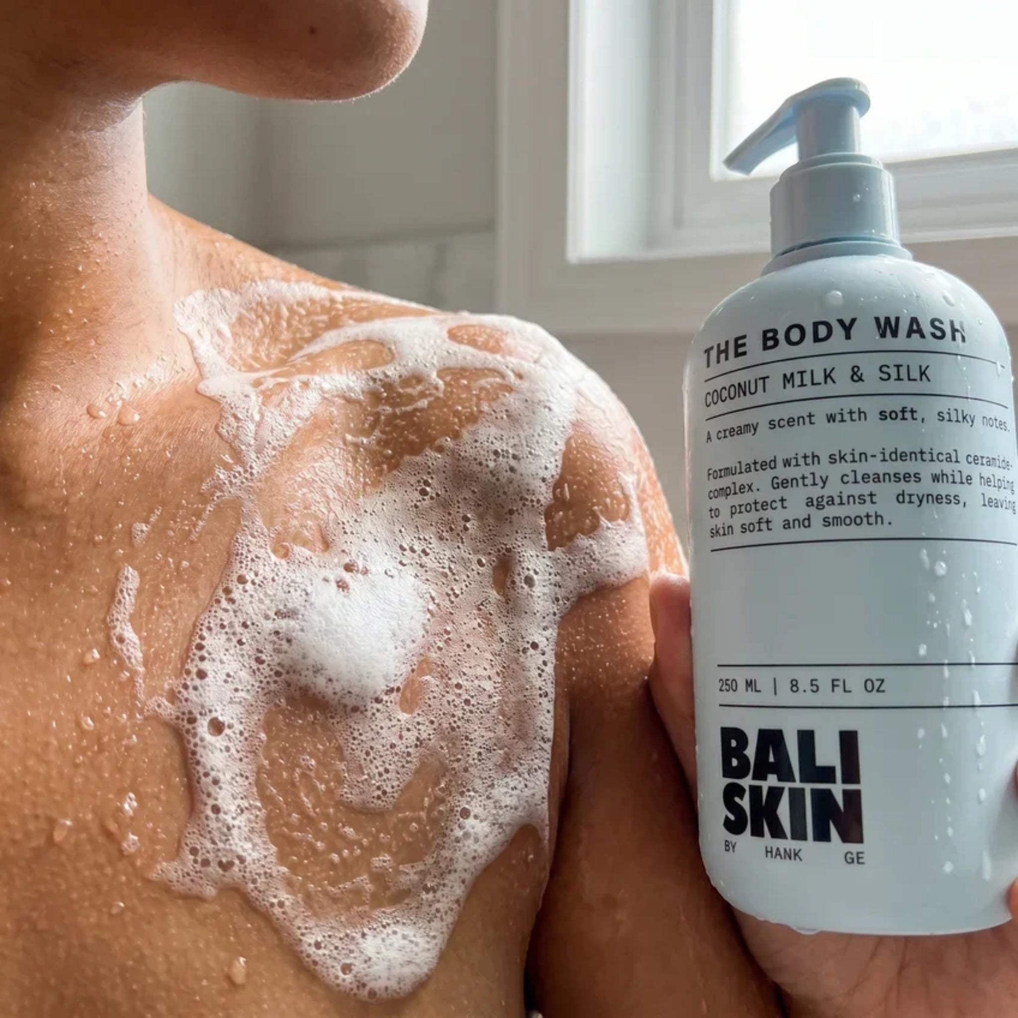 Skulder dækket af skum fra Bali Skin Body Wash Coconut Milk & Silk, pumpeflaske ved siden af i badeværelse.