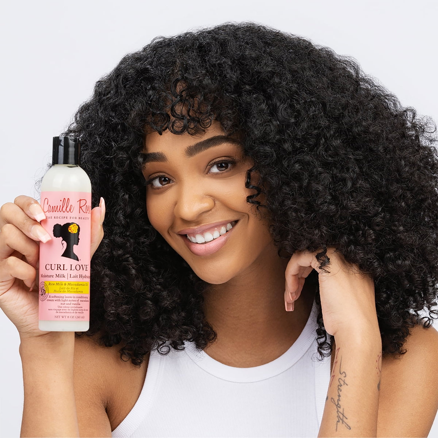 Camille Rose Curl Love Moisture Milk i hvid flaske med sort låg holdes af en kvinde med krøllet hår.