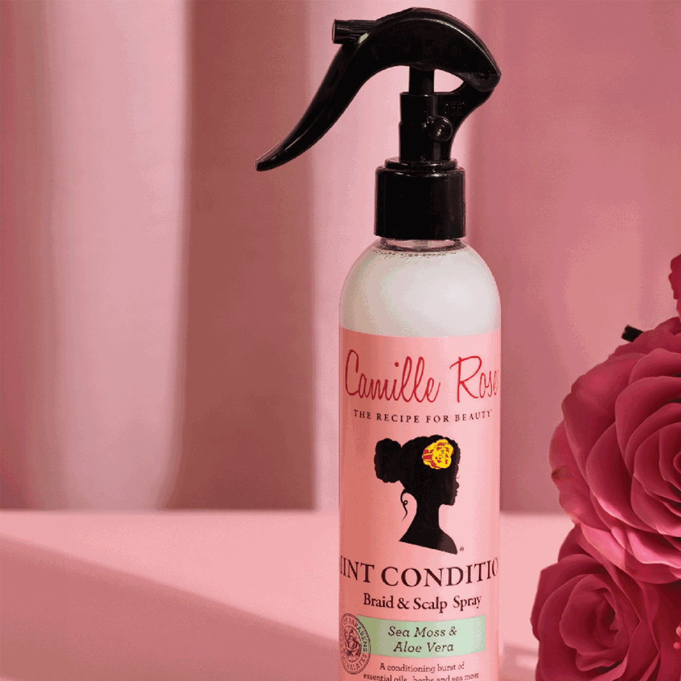 Pink sprayflaske med Camille Rose Mint Condition Braid & Scalp Spray. Produktet er afbildet mod en mørk baggrund sammen med kunstige rosa roser.