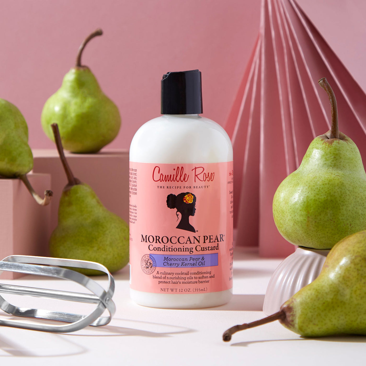 Camille Rose Moroccan Pear Conditioning Custard: Hvid flaske med lyserød etiket, omgivet af friske pærer og en pisker, på en lyserød baggrund.