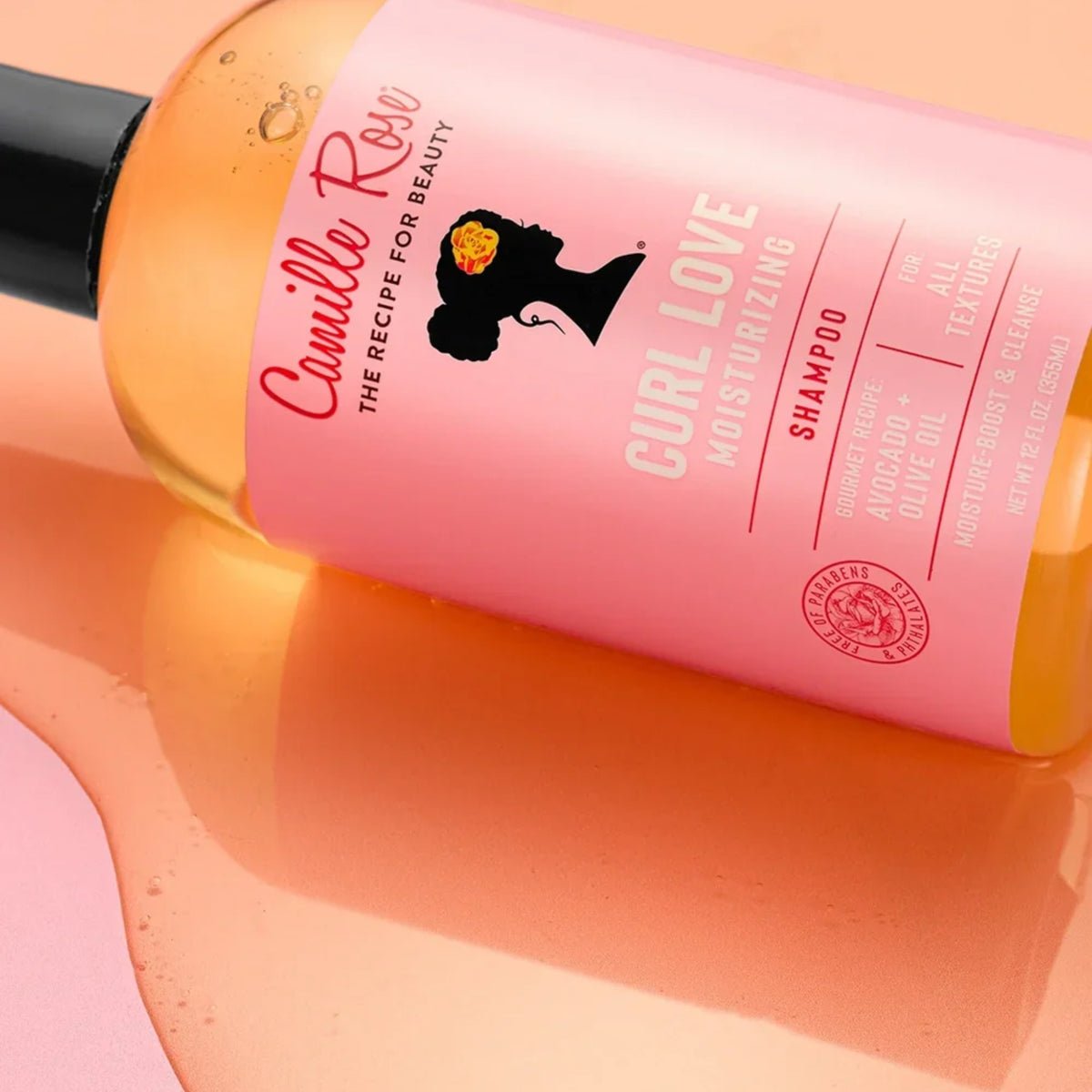 Camille Rose Curl Love Moisturizing Shampoo-flaske med pink etiket på lyserød baggrund med gyldne vandperler.