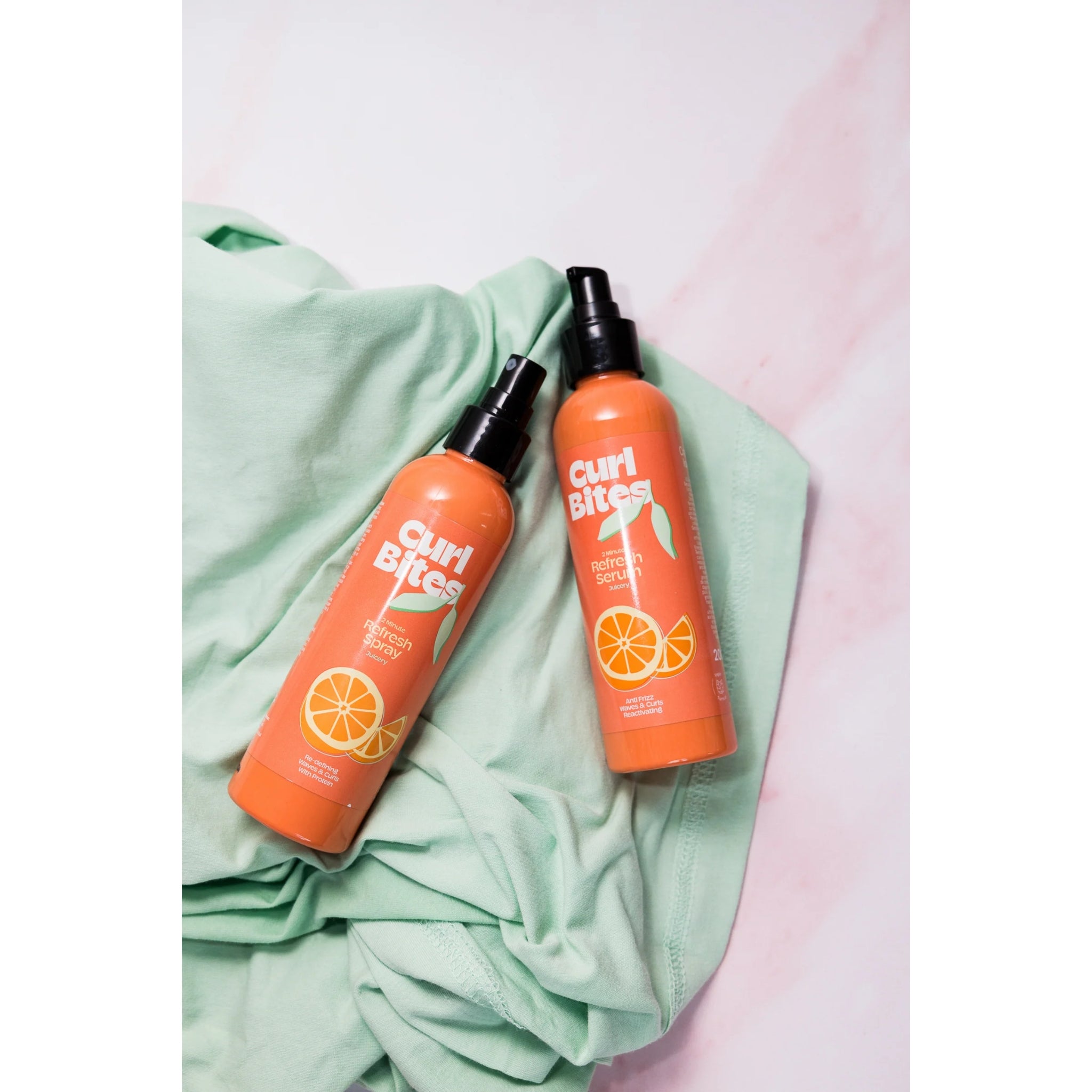 Curl Bites Refresh Spray og Refresh Serum i orange flasker med sort pumpe, placeret på mintgrøn stofbaggrund.