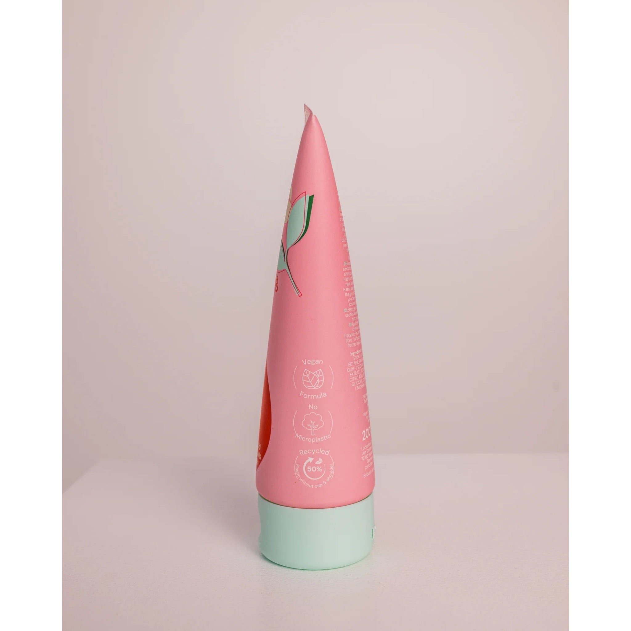 Konisk hårplejetube i lyserødt design med mintgrøn skruelåg, mærket med vegansk og mikroplastfri ikoner.