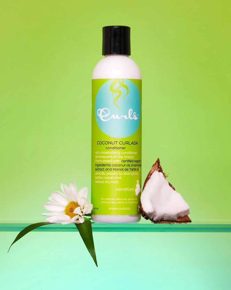 Curls Coconut Curlada Conditioner (240 ml) præsenteret med en kokosnød og en hvid blomst på en grøn baggrund. Hvid flaske med grøn etiket og sort låg. Balsam med kokosnød til krøllet hår.