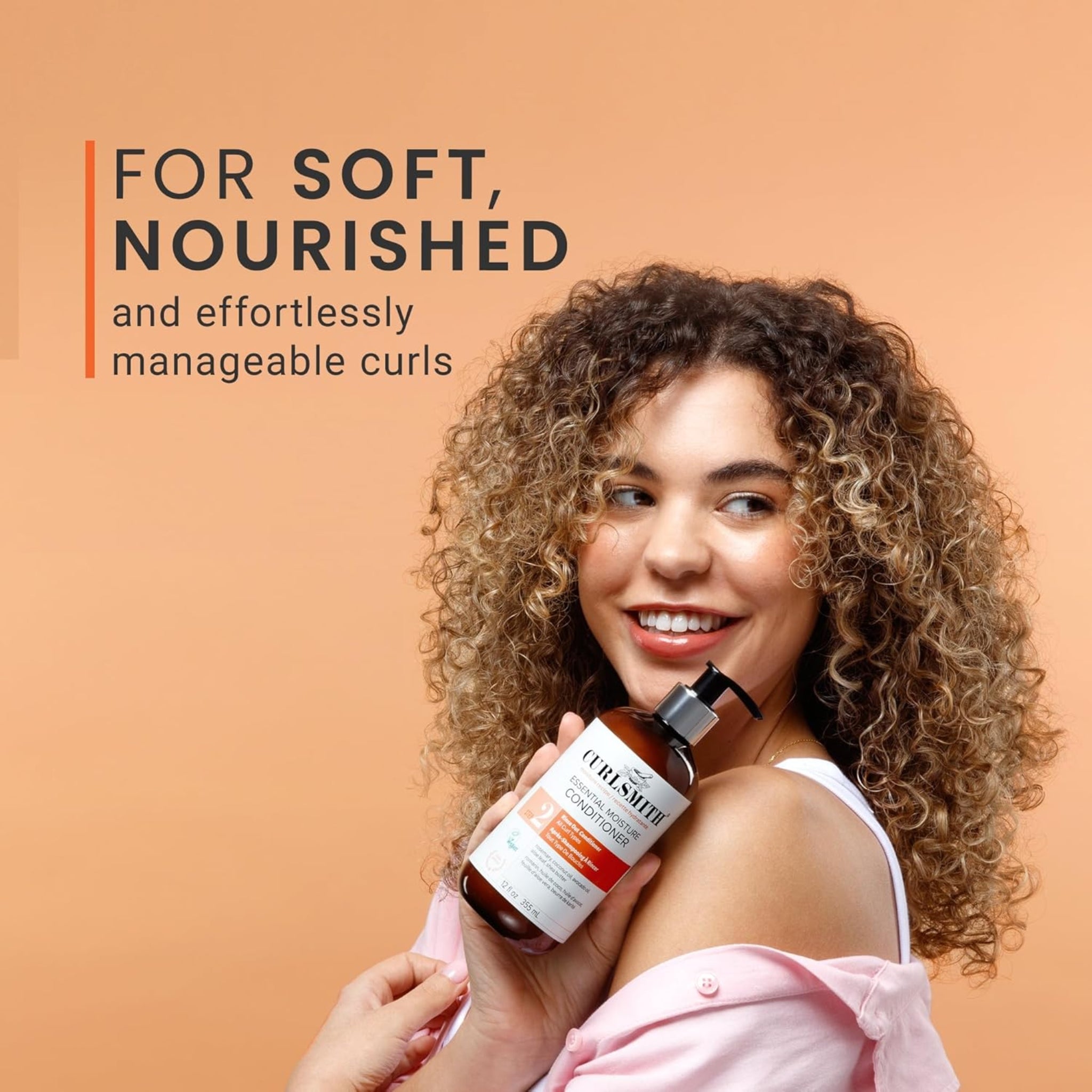 Kvinde med bløde, nærede krøller holder Curlsmith Essential Moisture Conditioner op mod skulderen og smiler.