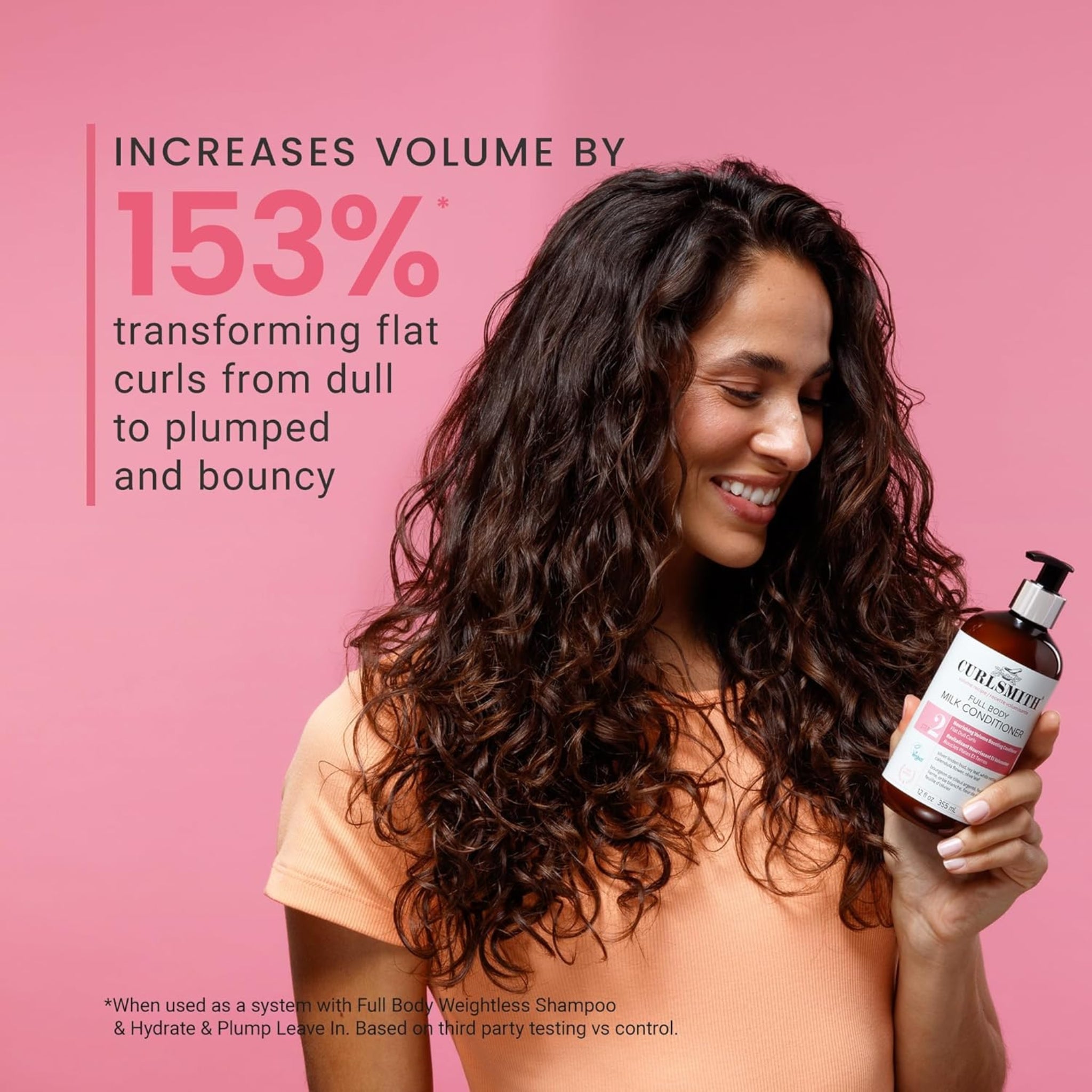 Kvinde med voluminøse, mørke krøller smiler og holder Curlsmith Full Body Milk Conditioner foran lyserød baggrund.