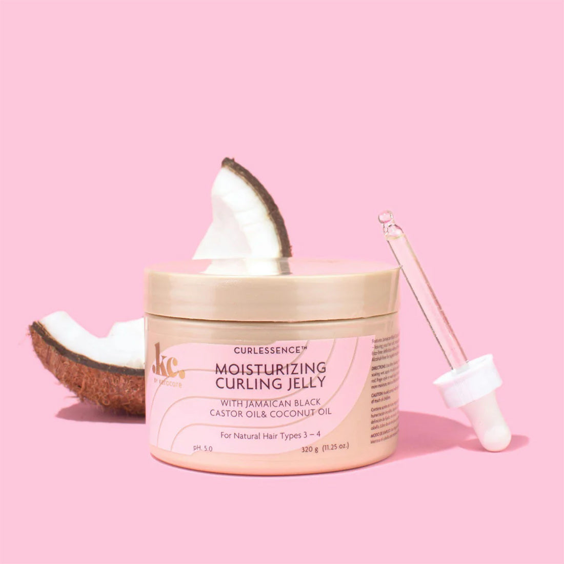 Beholder med KeraCare Curlessence Moisturizing Curling Jelly på en lyserød baggrund, ledsaget af stykker af en frisk kokosnød og en pipette. Fremhæver brugen af kokosolie i produktet.