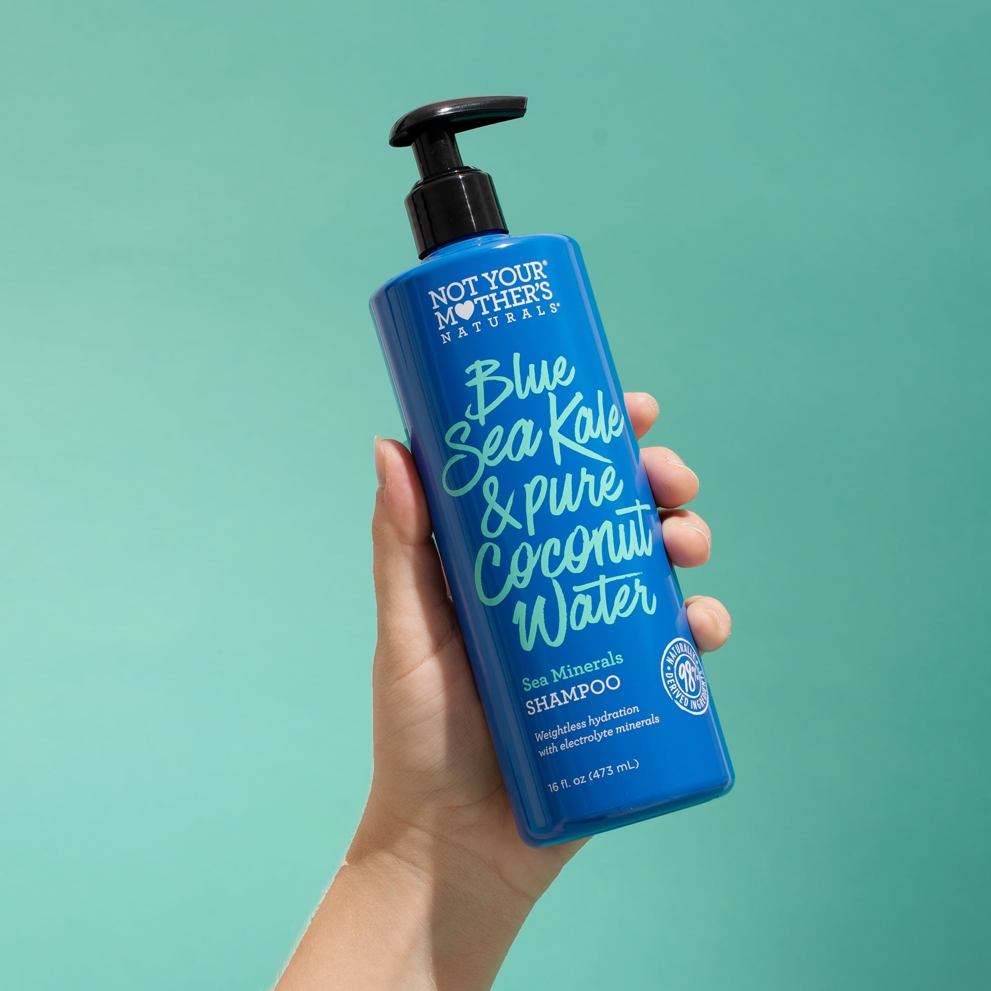 Nærbillede af en hånd, der holder en blå flaske Not Your Mother's Naturals Blue Sea Kale & Pure Coconut Water Sea Minerals Shampoo mod en turkis baggrund.