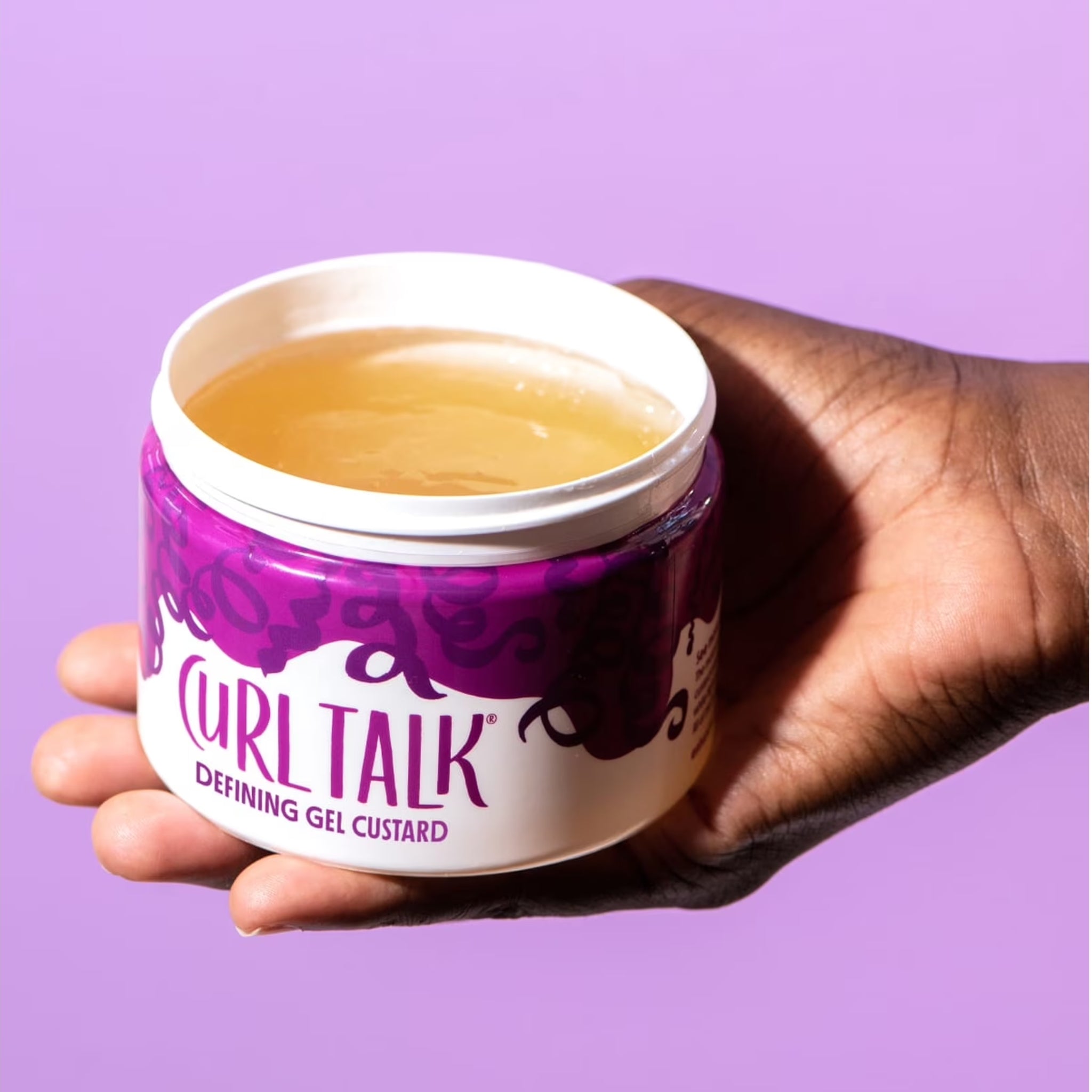 Hånd holder åben dåse med Curl Talk Defining Gel Custard, gylden gel synlig på lilla baggrund.