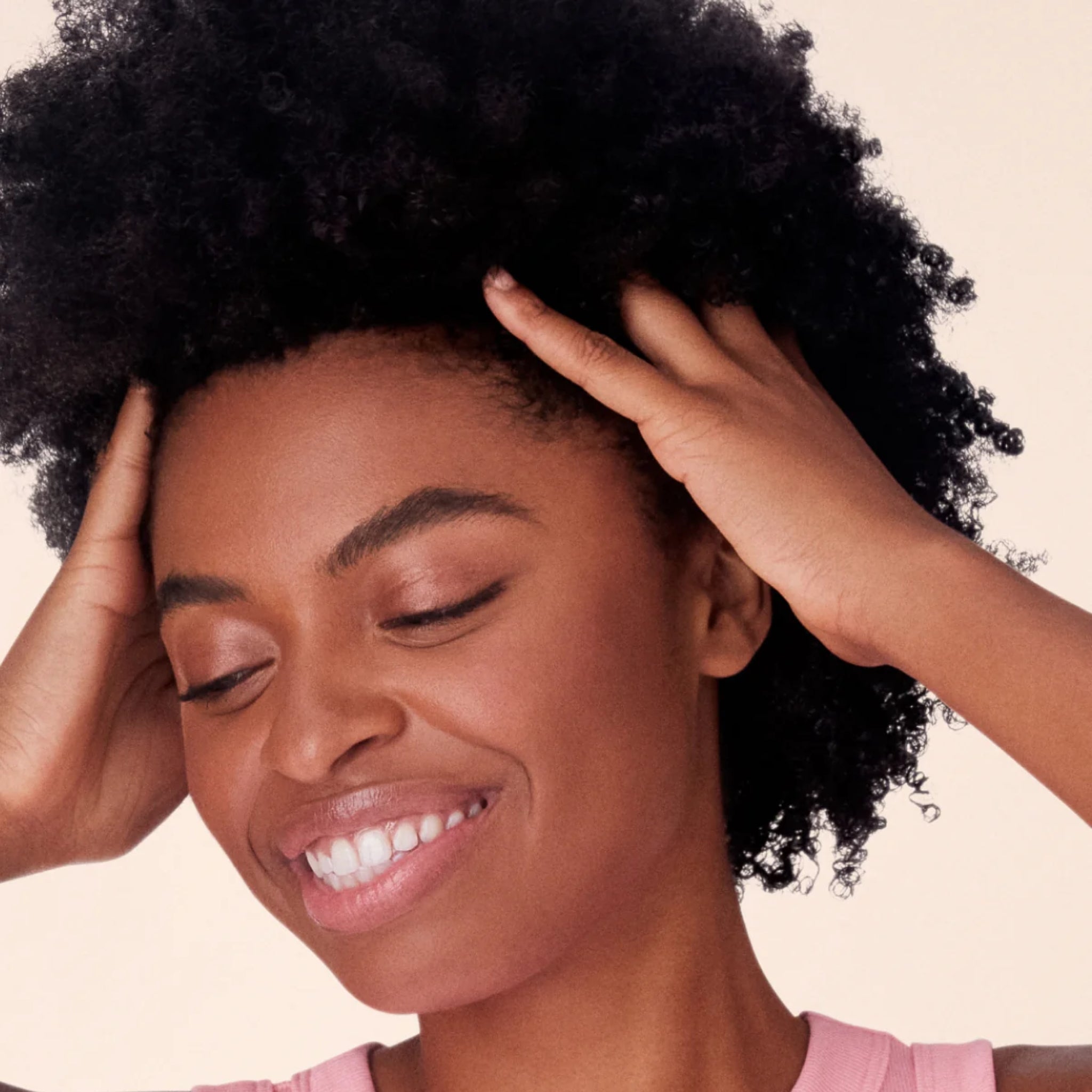 Smilende kvinde med naturligt afrohår, der rører ved sit krøllede hår med begge hænder mod beige baggrund.