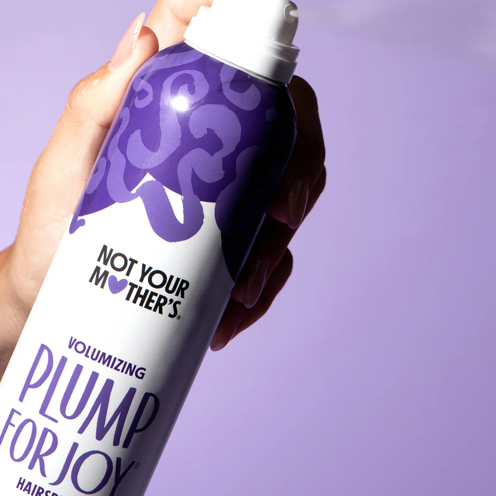 Hånd holder Not Your Mother's Plump For Joy volumizing hårspray i lilla og hvid dåse mod lilla baggrund.