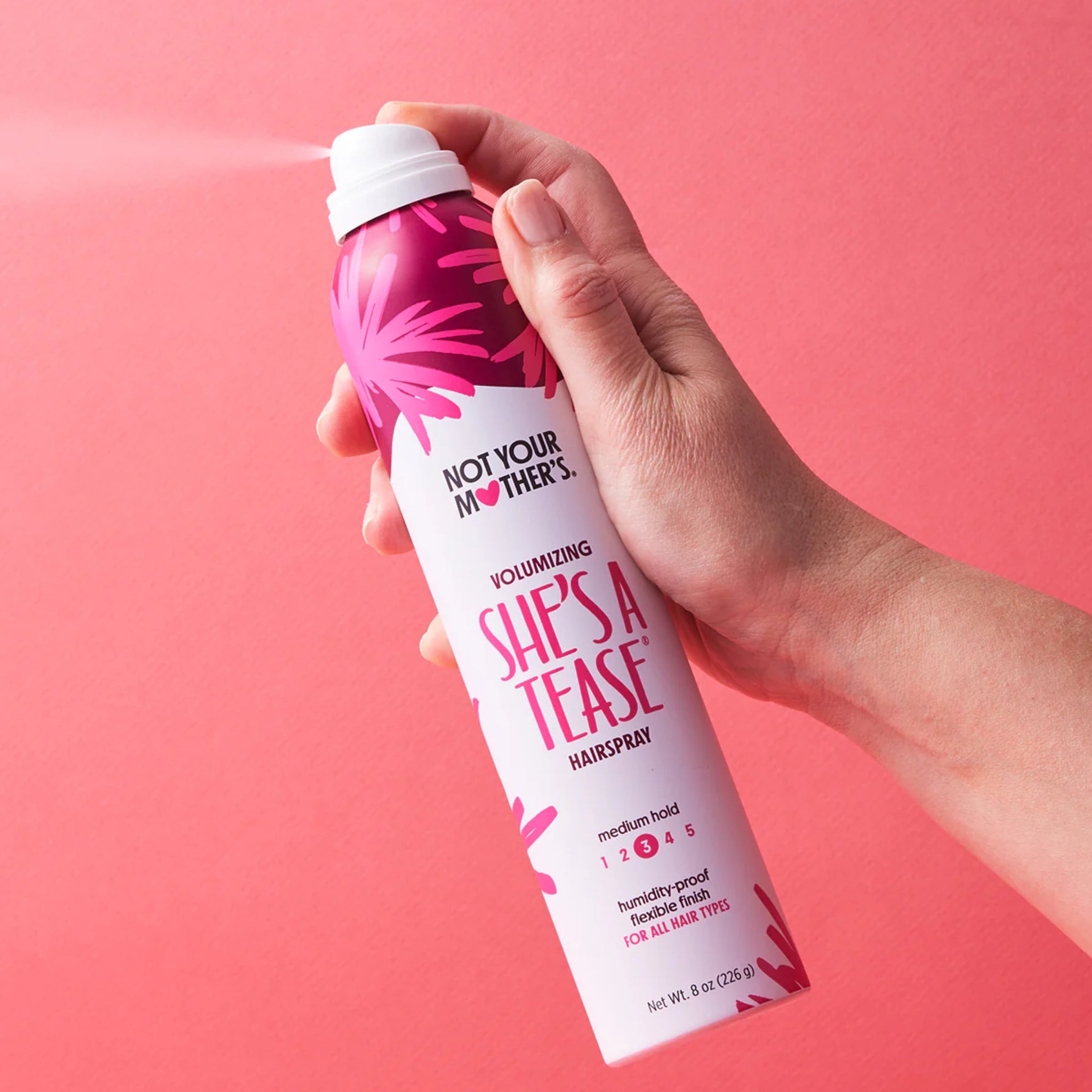 Hånd sprøjter Not Your Mother's She's A Tease volumizing hairspray mod lyserød baggrund.