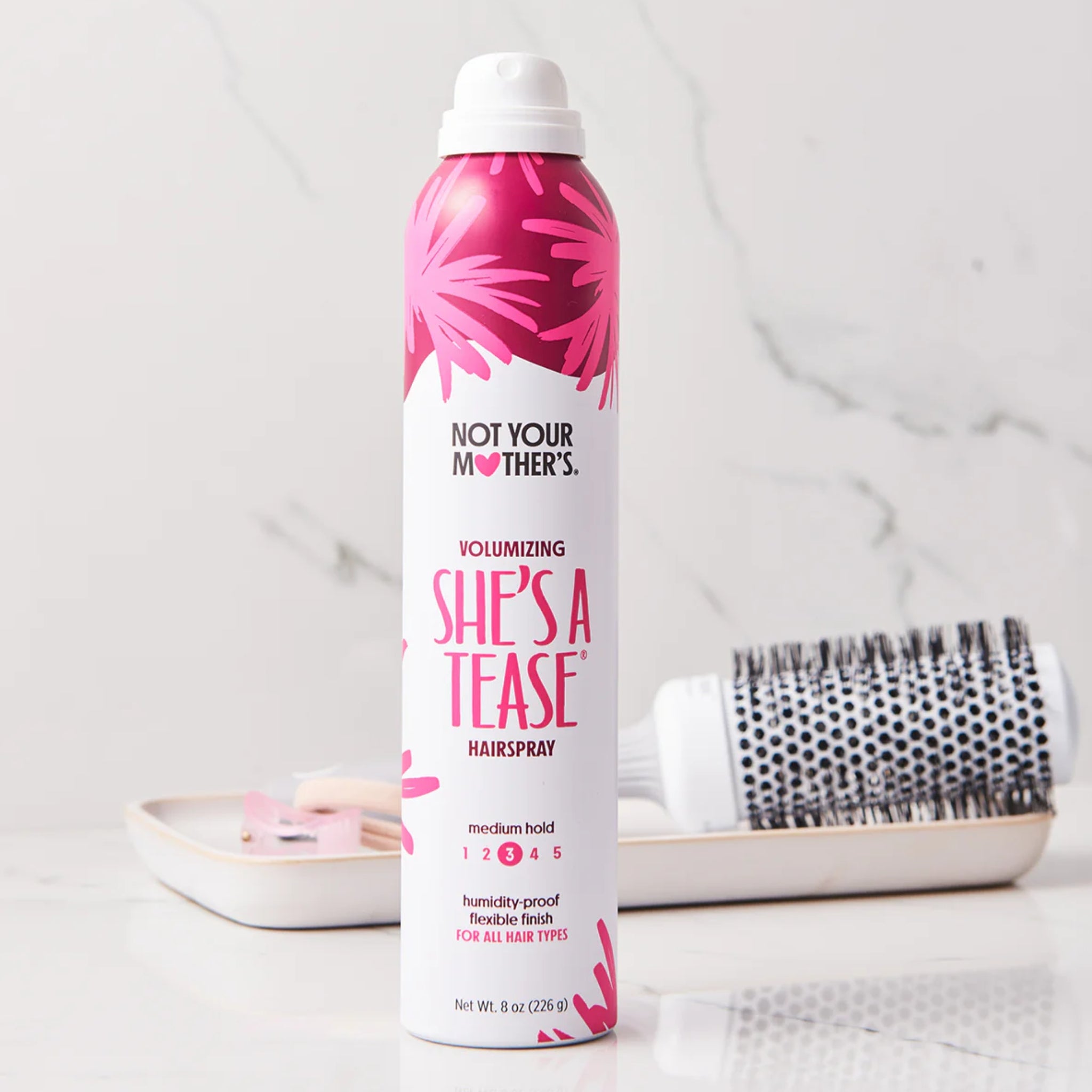 Not Your Mother's She's A Tease volumizing hairspray ved siden af rund børste på marmorbord.
