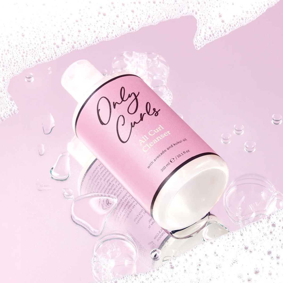 Only Curls All Curl Cleanser-flaske i lyserød emballage omgivet af skum og vandperler på lyslilla baggrund.