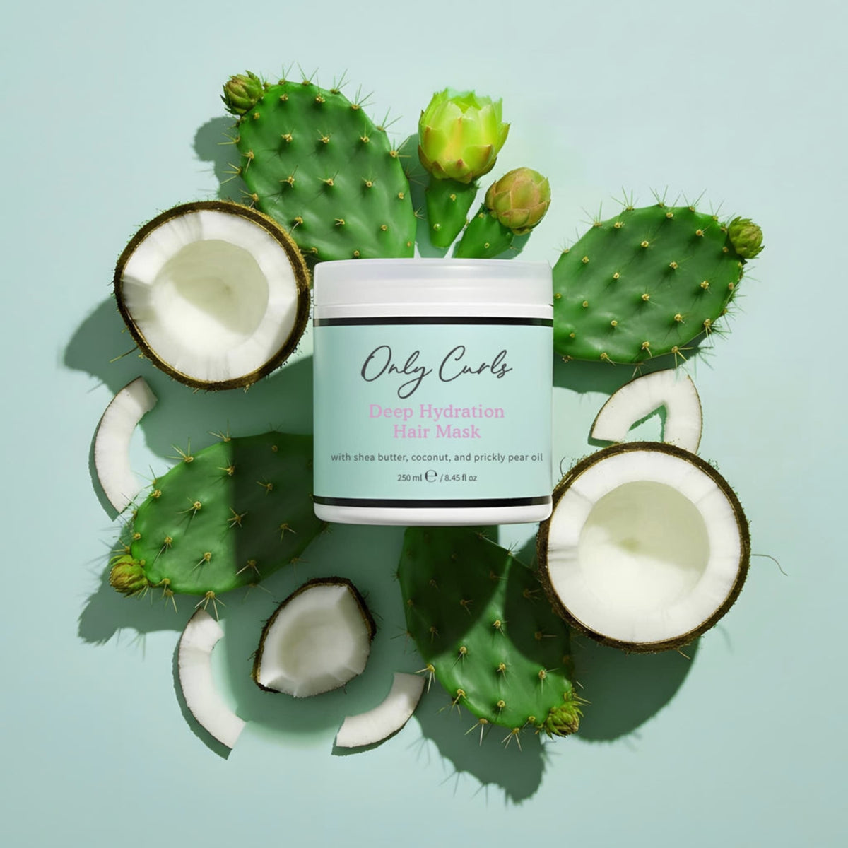 Only Curls Deep Hydration Hair Mask i hvid dåse omgivet af kokosnødder og grønne kaktusblade med blomster på mintgrøn baggrund.