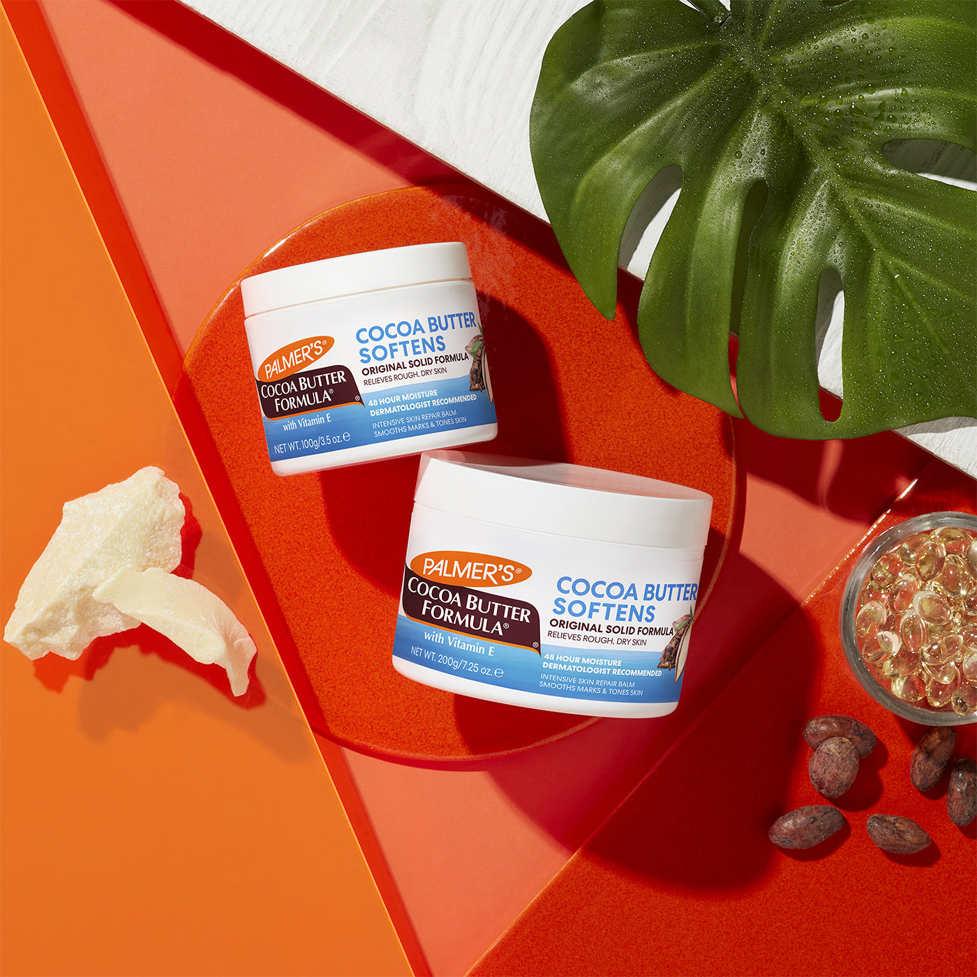 To beholdere af Palmers Cocoa Butter Formula Cream: en mindre 100g krukke og en større 200g krukke. Billedet viser også kakaosmør og E-vitamin kapsler.