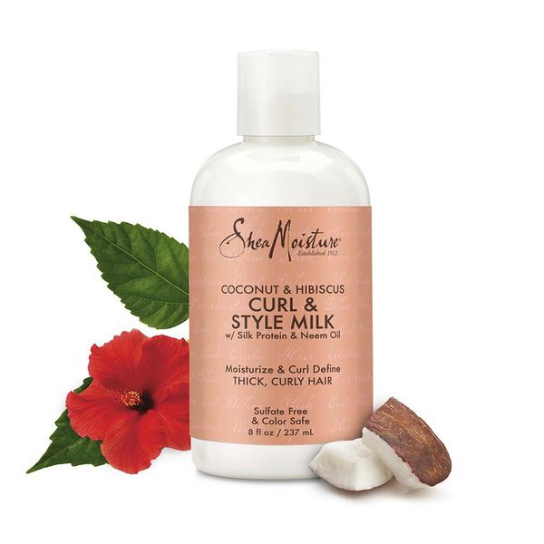 Hvid flaske Shea Moisture Coconut & Hibiscus Curl & Style Milk (237 ml) placeret ved siden af en rød hibiscusblomst med grønne blade og et stykke af en kokosnød.