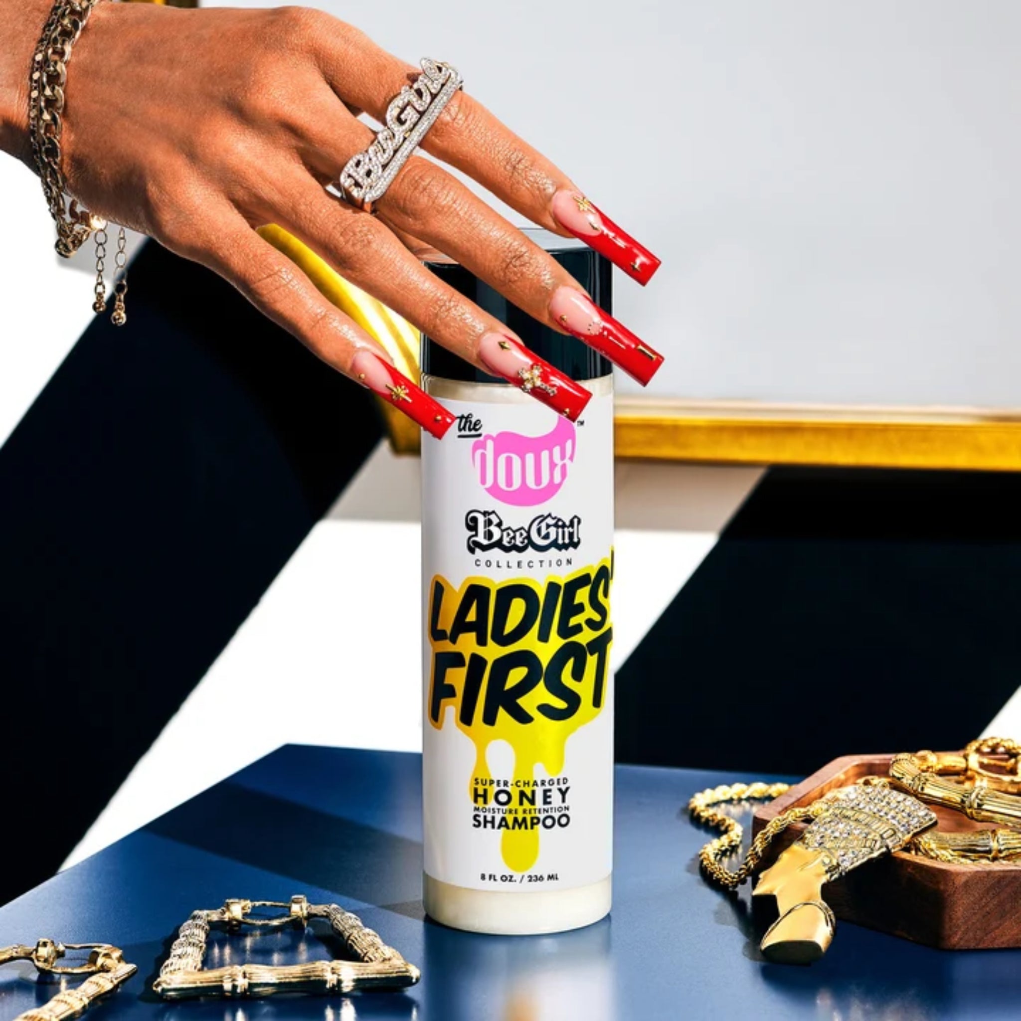 Hånd med lange røde negle rører The Doux Bee Girl Ladies First honning-shampoo ved siden af guldsmykker.
