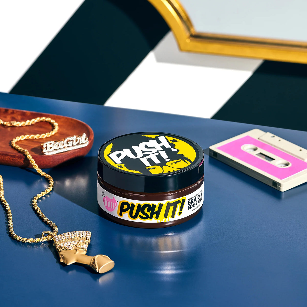 Doux Push It! Braid & Edge Gel i sort/gul krukke på blåt bord med guldkæde og kassettebånd.